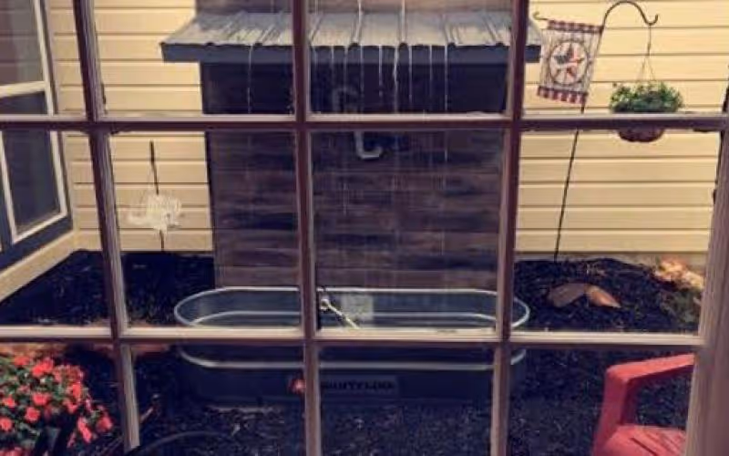 View through a window showing a small outdoor garden area with a wooden shed, a metal trough, a hanging planter with greenery, a decorative flag, and a red chair. There are flowers planted in the garden bed and mulch covering the ground.