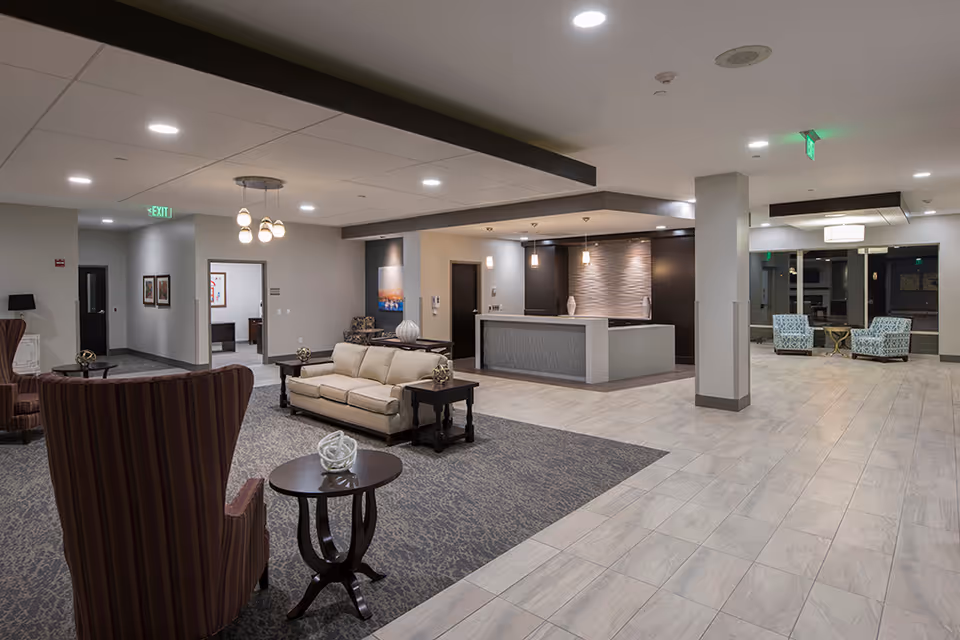A spacious and modern senior living facility common area with a reception desk, comfortable seating including a beige sofa and striped armchairs, small tables with decorative items, and soft overhead lighting. The floor is a combination of carpet and tile, and there are exit signs and doors visible in the background.