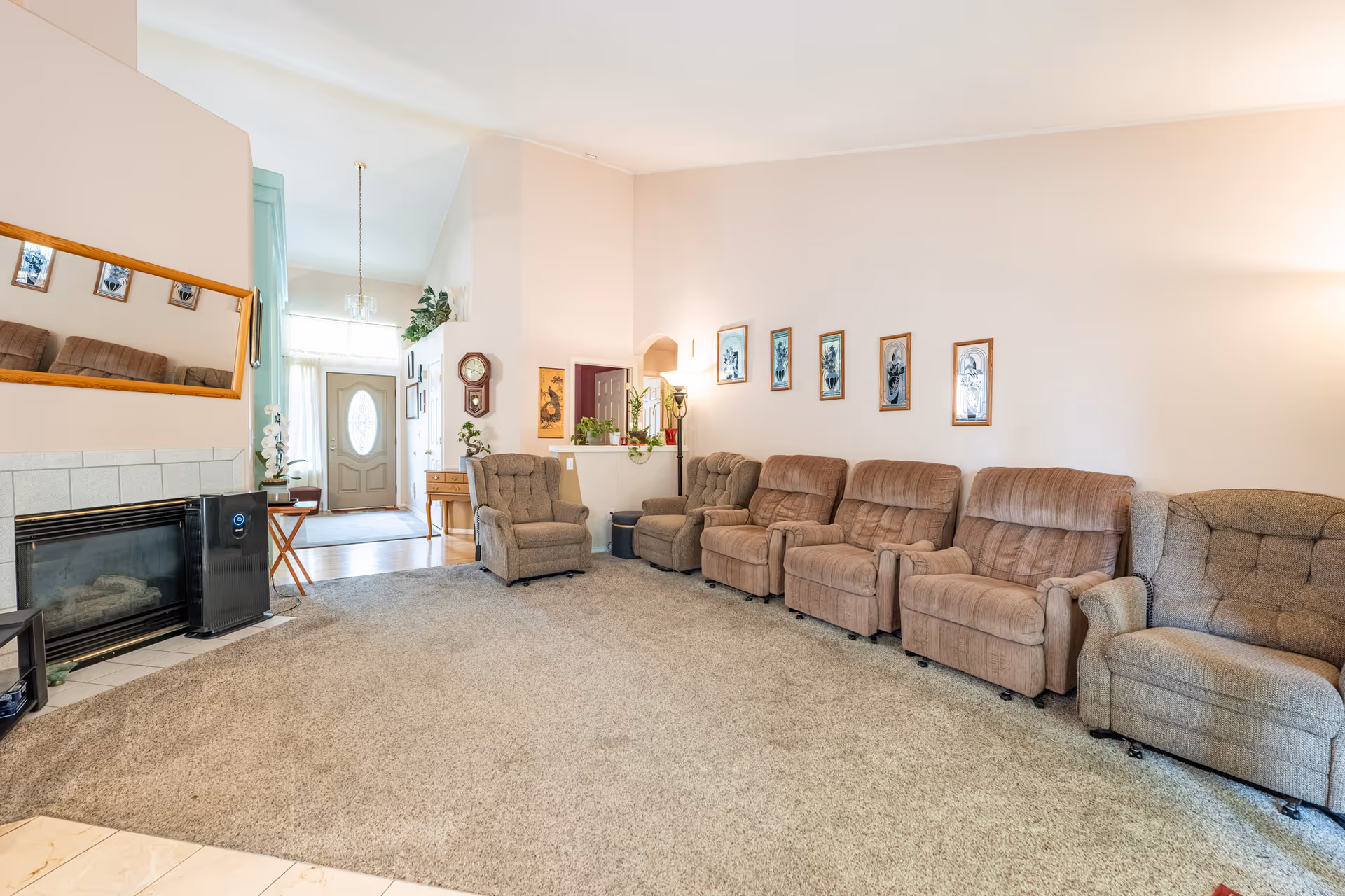 A spacious living room with a row of six cushioned recliner chairs lined up against a beige wall adorned with framed pictures. The room features a carpeted floor, a fireplace with a large mirror above it, and an air purifier next to the fireplace. The entrance door with an oval window is visible in the background, along with a small table and some plants.
