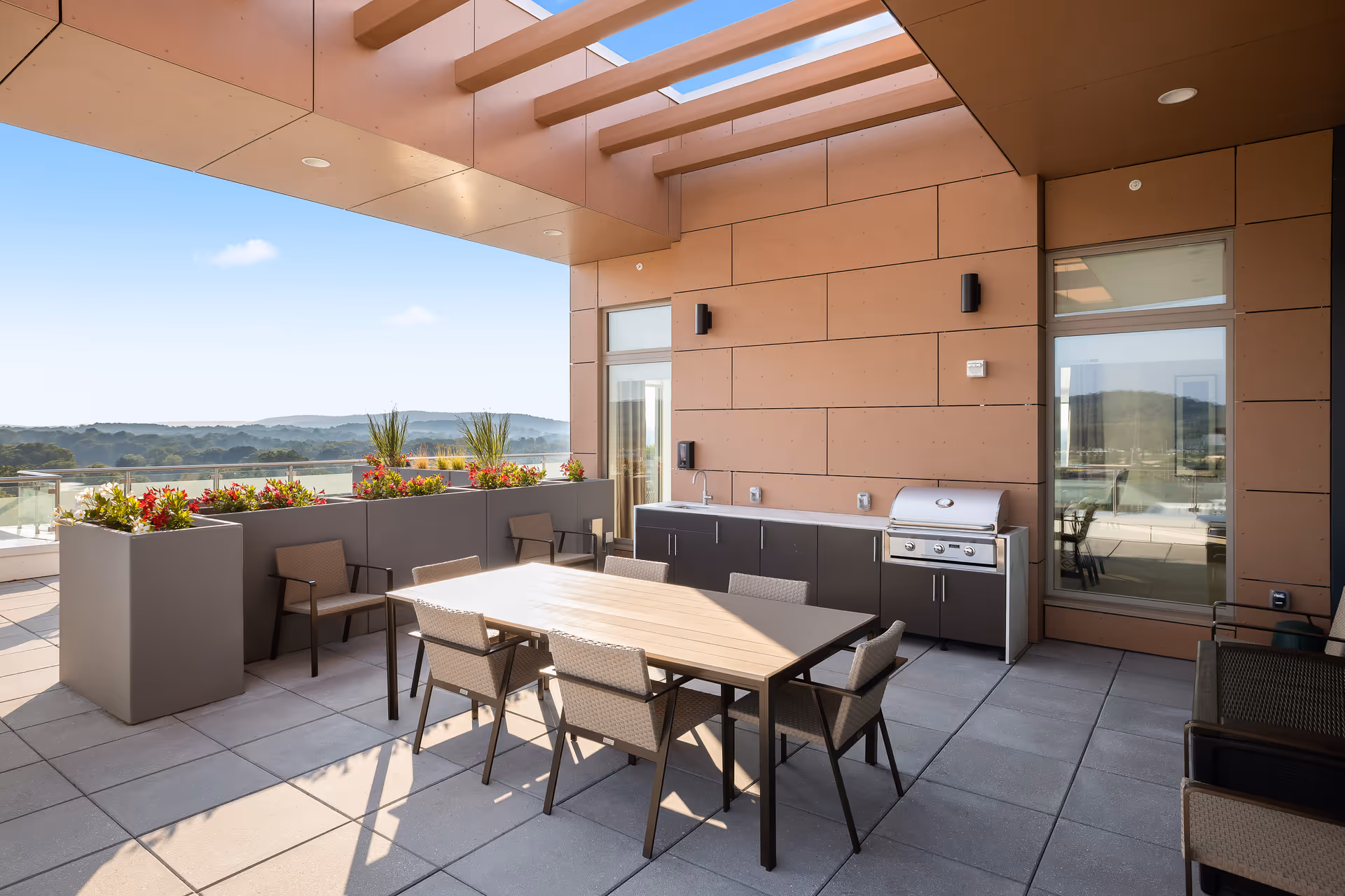 Outdoor patio area with a dining table and six chairs, a built-in grill and sink, large planters with flowers, and a view of distant hills under a clear blue sky.
