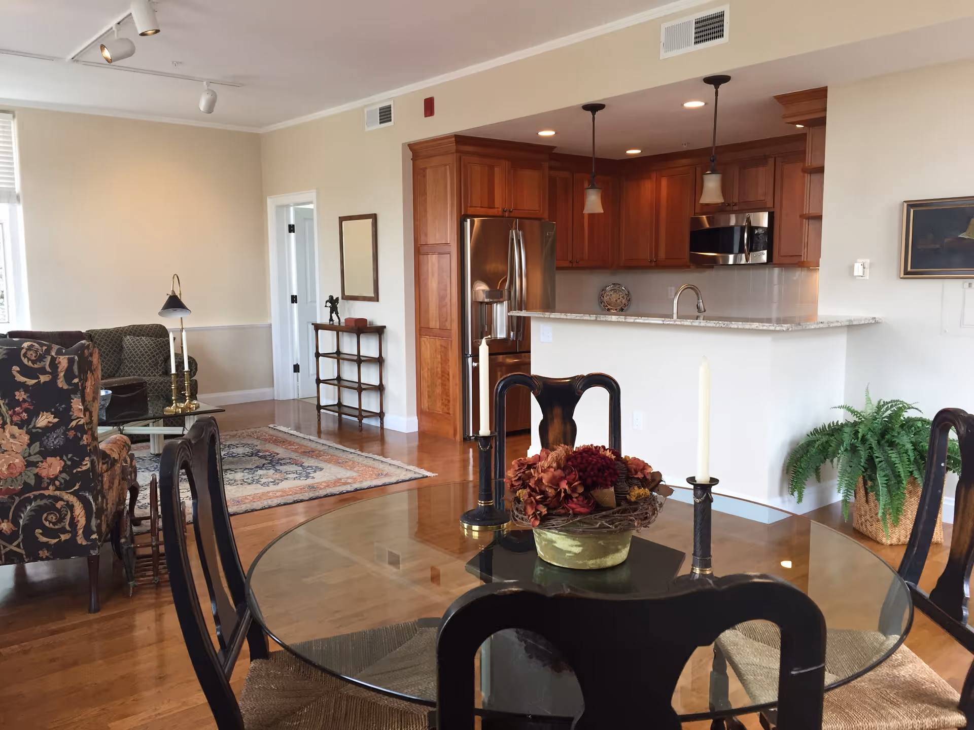 Interior view of a senior living facility showing a dining area with a round glass table and four chairs, a centerpiece with flowers and two candles on the table. In the background, there is a kitchen with wooden cabinets, a stainless steel refrigerator, a microwave, and a granite countertop with pendant lights. To the left, there is a living area with floral and green upholstered chairs, a glass coffee table, a floor lamp, and a patterned rug on wooden flooring. A small shelf with decorative items and a mirror is visible against the wall.