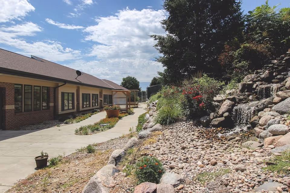 Outdoor view of Independence Houses of Northview showing a paved walkway alongside a building with large windows. On the right side, there is a landscaped area with rocks, plants, flowers, and a small cascading waterfall. The sky is partly cloudy.