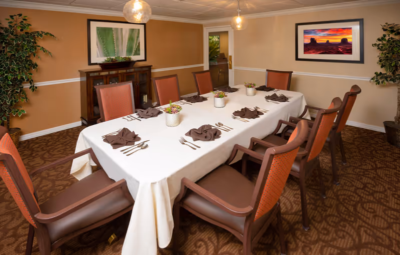 A dining room with a long rectangular table covered with a white tablecloth, set with brown folded napkins, silverware, and small potted plants as centerpieces. The room has eight wooden chairs with brown cushions and orange backs. The walls are beige with white trim, decorated with two framed pictures and two potted plants in the corners. Two pendant lights hang from the ceiling.