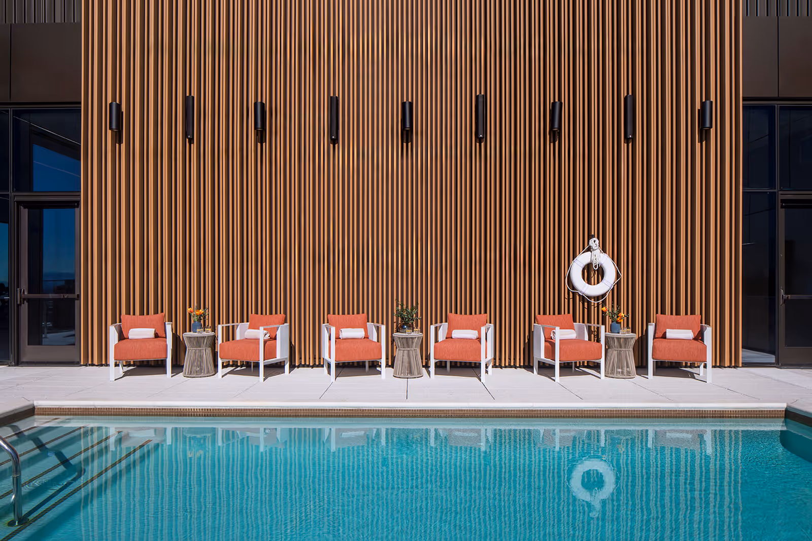 Poolside scene with six orange cushioned chairs and side tables lined against a tall wooden slatted wall with a life ring, reflected in the swimming pool.