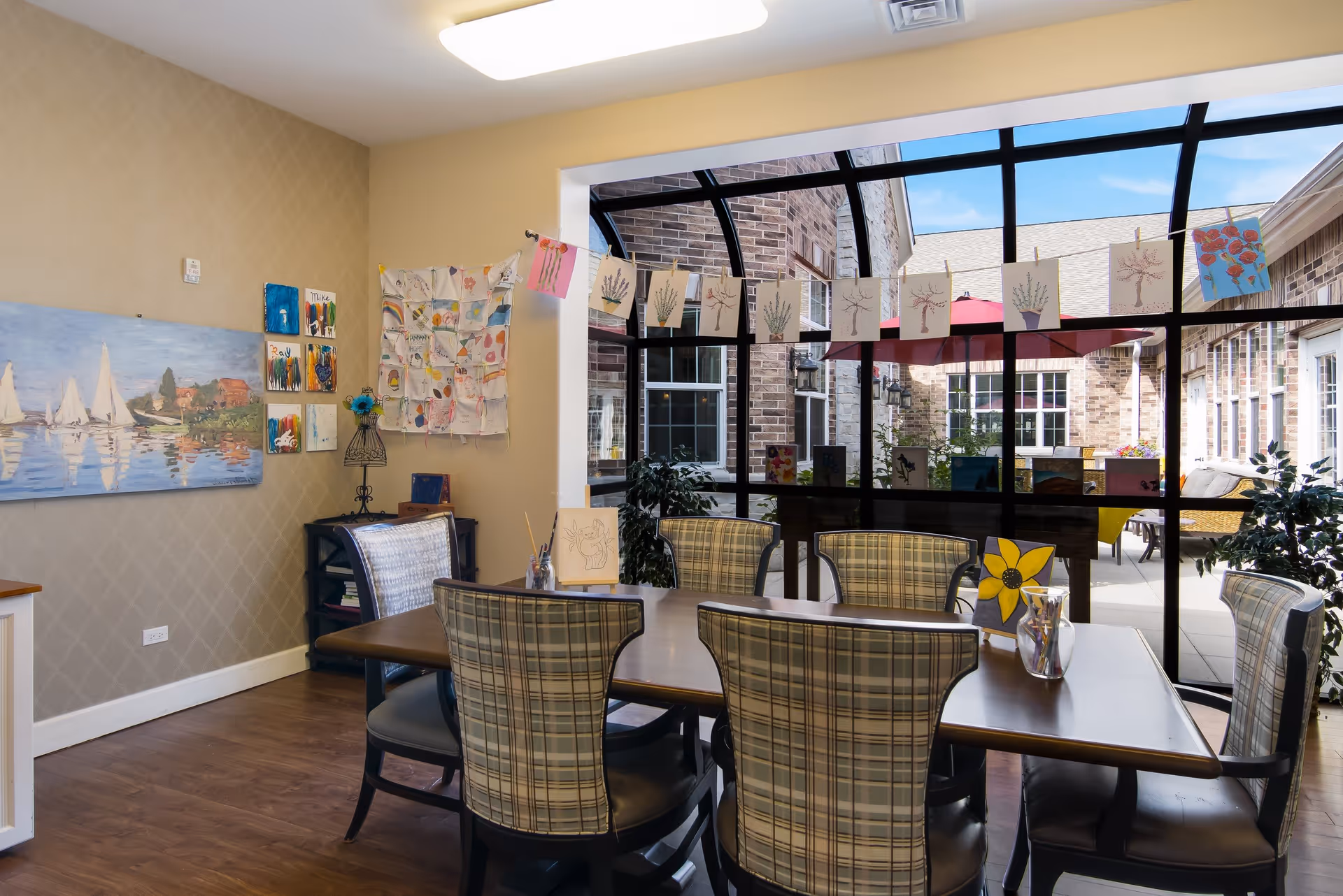 A cozy indoor dining area with a wooden table surrounded by six plaid upholstered chairs. The room has a large window wall looking out onto a courtyard with brick buildings and red umbrellas. Various colorful artworks and paintings are displayed on the walls and hanging on a string across the window. The floor is wooden, and the room is well-lit with a ceiling light.