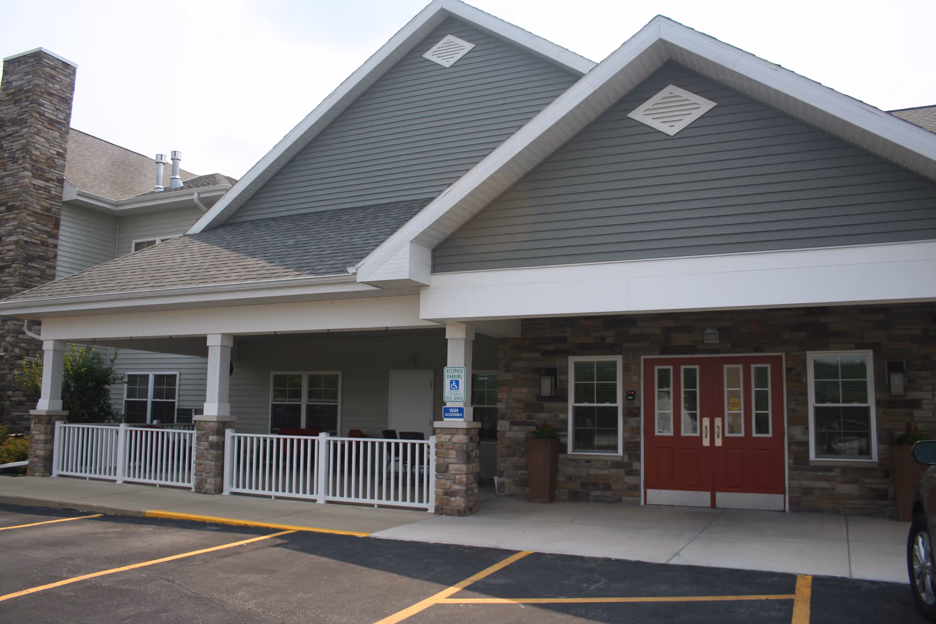 Exterior view of the entrance to Milestone Senior Living Cross Plains featuring a covered porch with white railings, stone pillars, and red double doors. There are windows on either side of the doors and a reserved parking space for handicapped access in front.