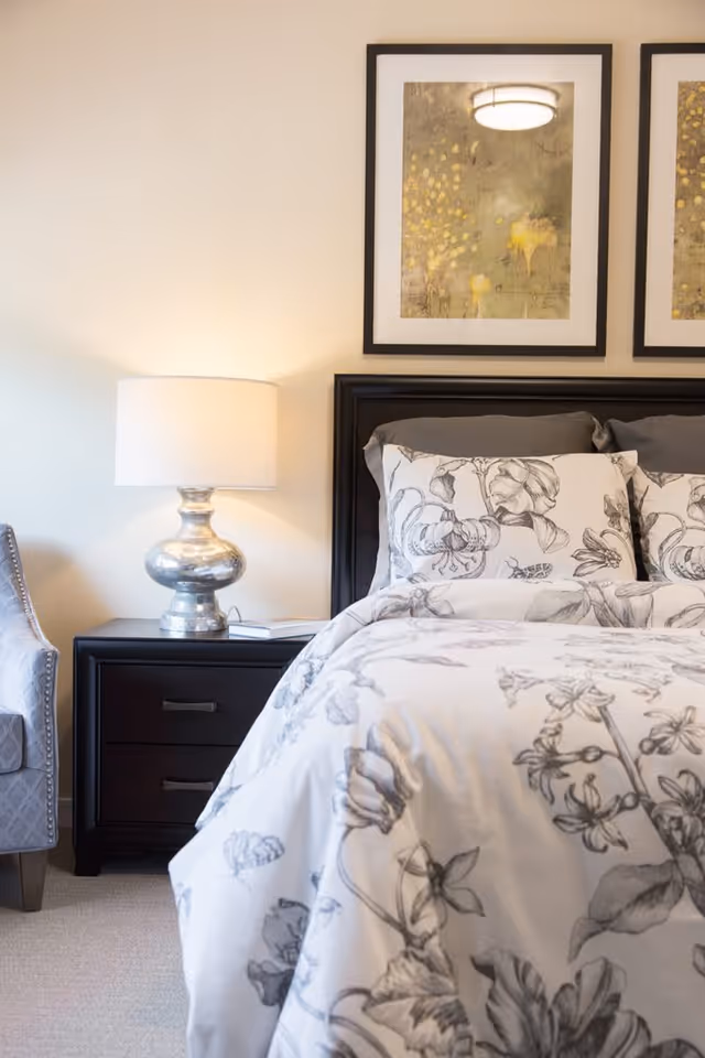 A cozy bedroom featuring a bed with floral patterned bedding and pillows, a dark wooden headboard, a matching nightstand with two drawers, a silver table lamp with a white lampshade, and a gray upholstered chair. Two framed abstract artworks hang on the wall above the bed.