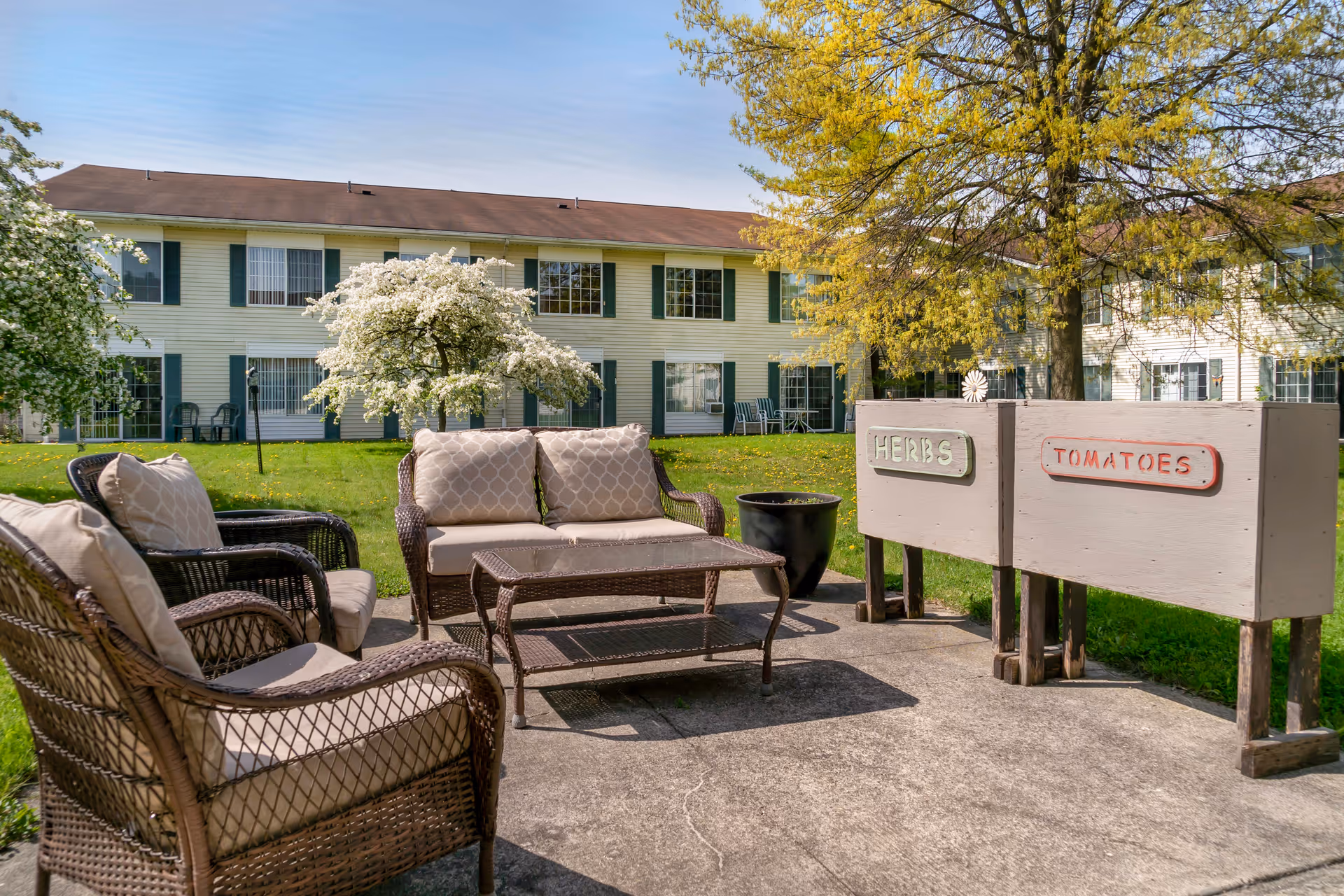 Outdoor patio area with wicker furniture including a loveseat, two armchairs, and a coffee table. Behind the seating area are two raised garden beds labeled 'HERBS' and 'TOMATOES'. In the background, there is a two-story building with green shutters and several windows, along with trees and a grassy lawn.