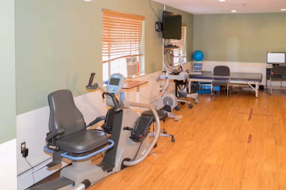 Rehabilitation exercise room with recumbent exercise bikes, therapy tables, chairs and an exercise ball on a wood floor.