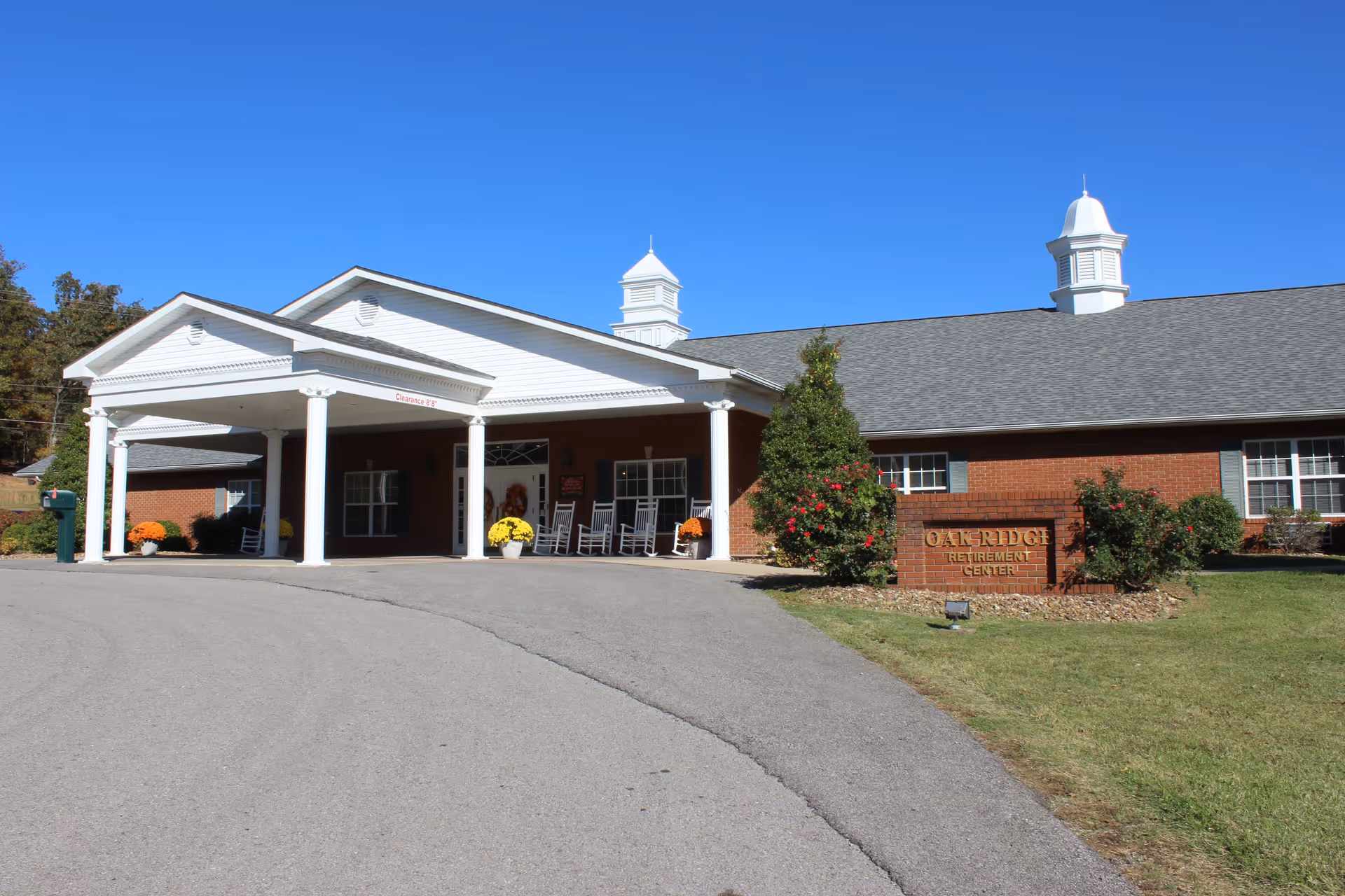 Front exterior view of Oak Ridge Retirement Center building with a covered entrance supported by white columns, rocking chairs on the porch, and a sign in front surrounded by bushes and flowers under a clear blue sky.