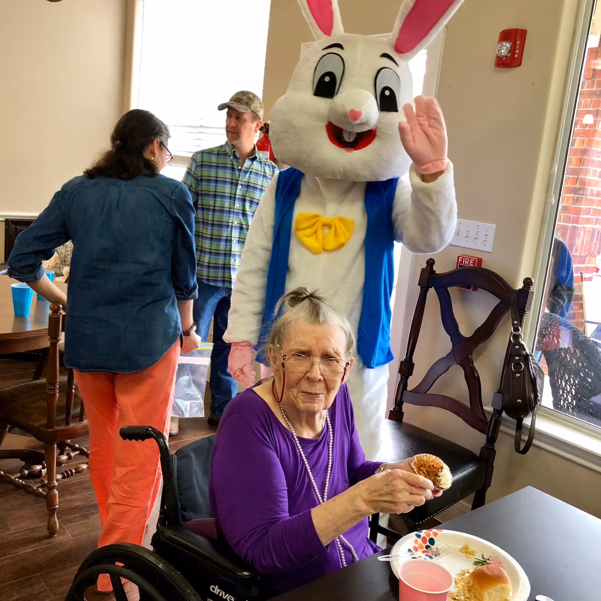 An elderly woman in a wheelchair eating a cupcake at a table during a gathering with a person in an Easter bunny costume waving behind her and other people nearby.