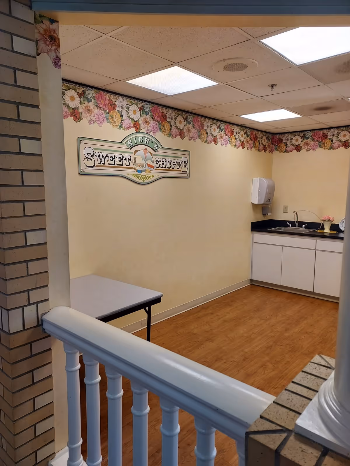 Interior view of a small room with a wooden floor, a white railing in the foreground, a brick column on the left, and a counter with a sink and soap dispenser on the right. The walls are decorated with a floral border near the ceiling and a sign that reads 'Nick's Sweet Shoppe'.