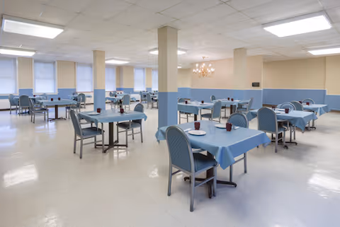 A spacious dining room with multiple tables covered in blue tablecloths, each surrounded by blue cushioned chairs. The room has beige and light blue walls, several support columns, large windows with blinds, and bright overhead lighting. A chandelier is visible on one wall.