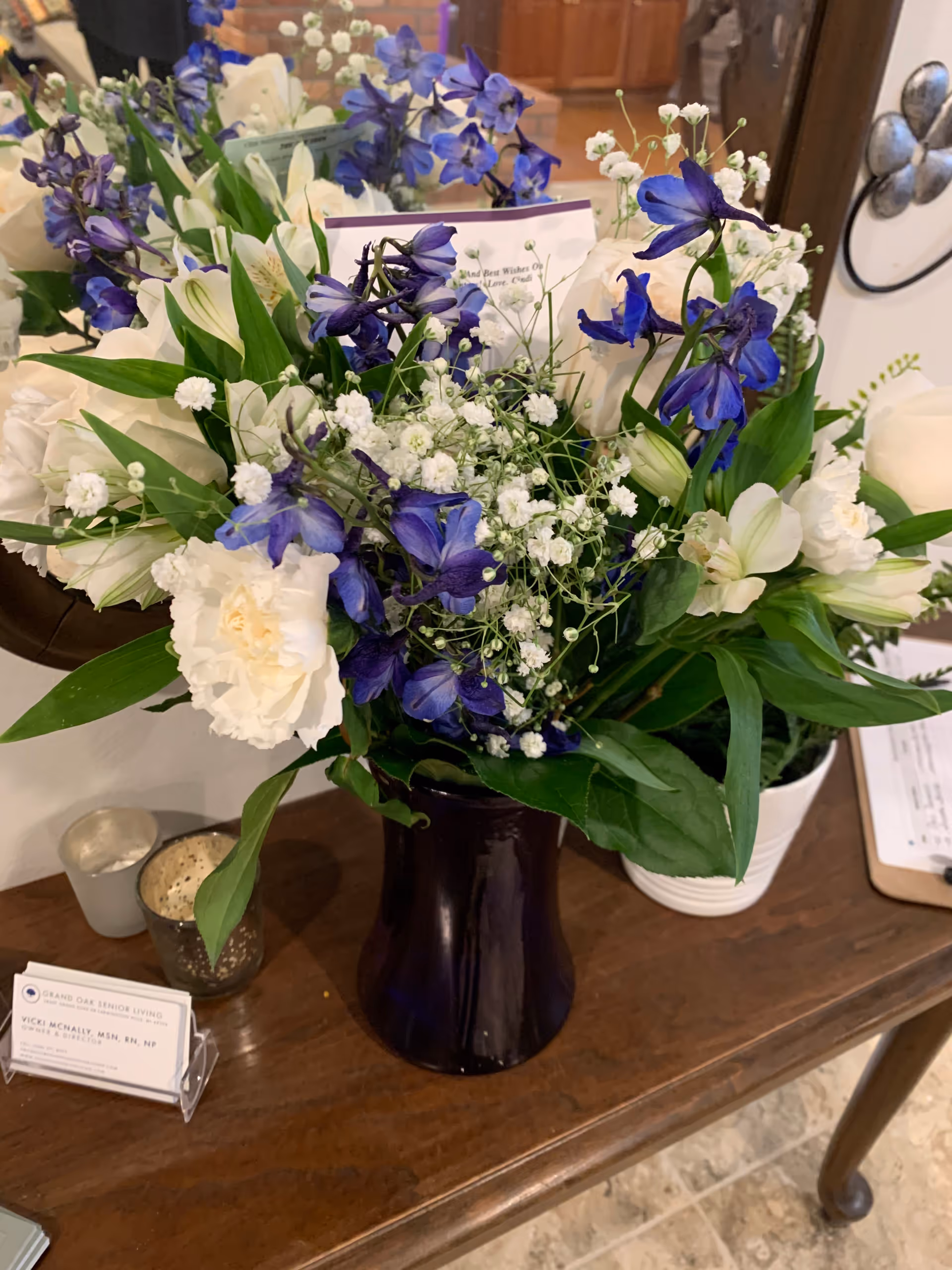 A bouquet of white and purple flowers arranged in a dark vase on a wooden table. Behind the bouquet, there is a mirror reflecting part of the flowers and a brick wall. On the table, there are two small candle holders and a business card holder with a card for Vicki McNally, Director at Grand Oak Senior Living.