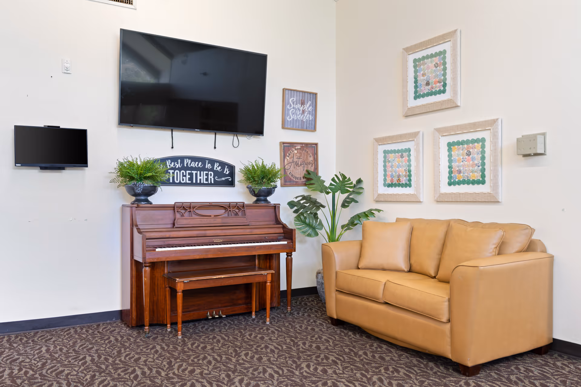 A cozy interior corner featuring a wooden upright piano with a matching bench, two potted plants on top, and a wall-mounted flat-screen TV above it. To the right, there is a tan leather loveseat with three cushions, a large green leafy plant in a pot, and three framed quilt-like artworks on the wall. The carpet has a patterned design.