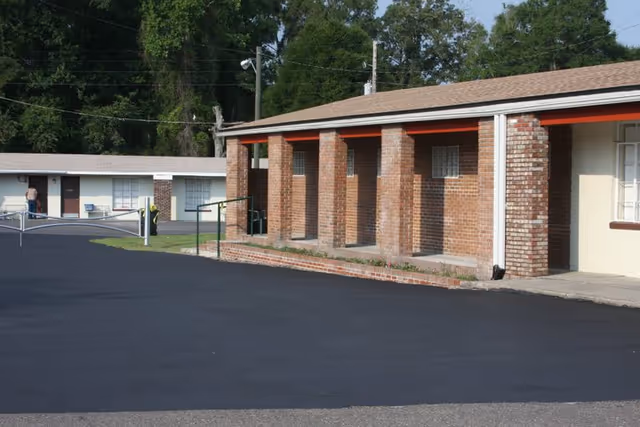 Exterior view of a single-story brick building with a covered porch supported by brick columns. The building is part of a complex with additional single-story structures visible in the background, surrounded by trees and a paved driveway or parking area in the foreground.
