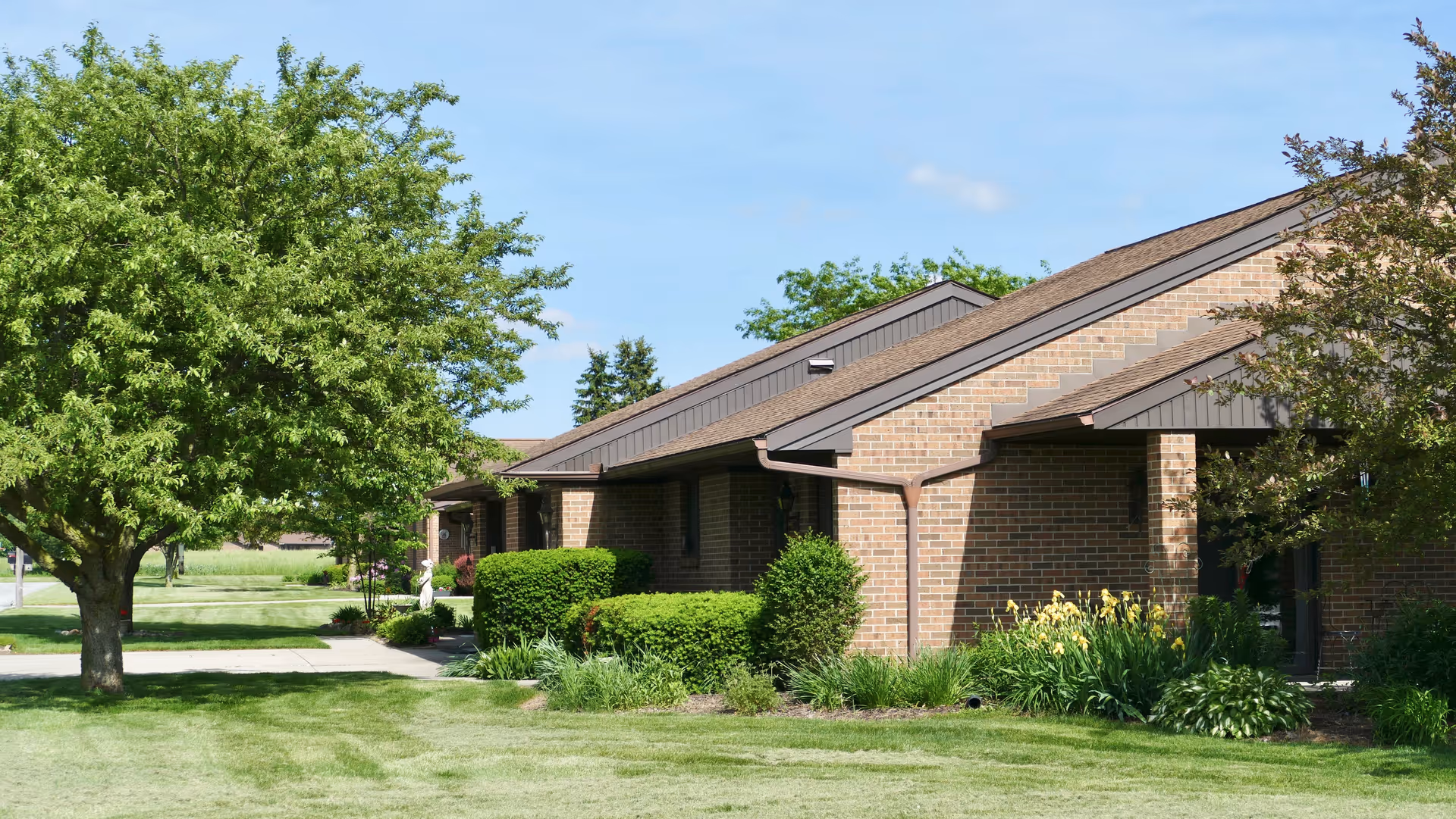 Single-story brick building with a manicured lawn, shrubs and a large tree under a blue sky.