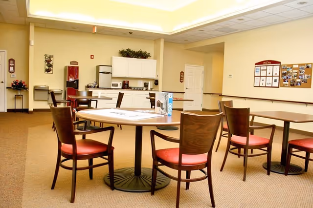 A well-lit common area with several round wooden tables and chairs with red cushions. In the background, there is a kitchenette with white cabinets, a microwave, coffee maker, and a refrigerator. The walls are light-colored, and there are bulletin boards and decorations on the walls.