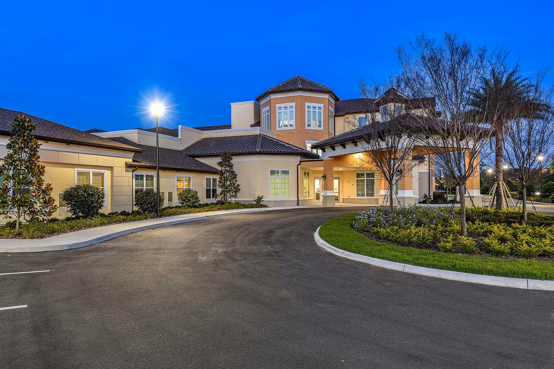 Exterior view of Providence Living at Hunter's Creek facility at dusk, showing a well-lit entrance with a covered drop-off area, landscaped greenery, trees, and a curved driveway.