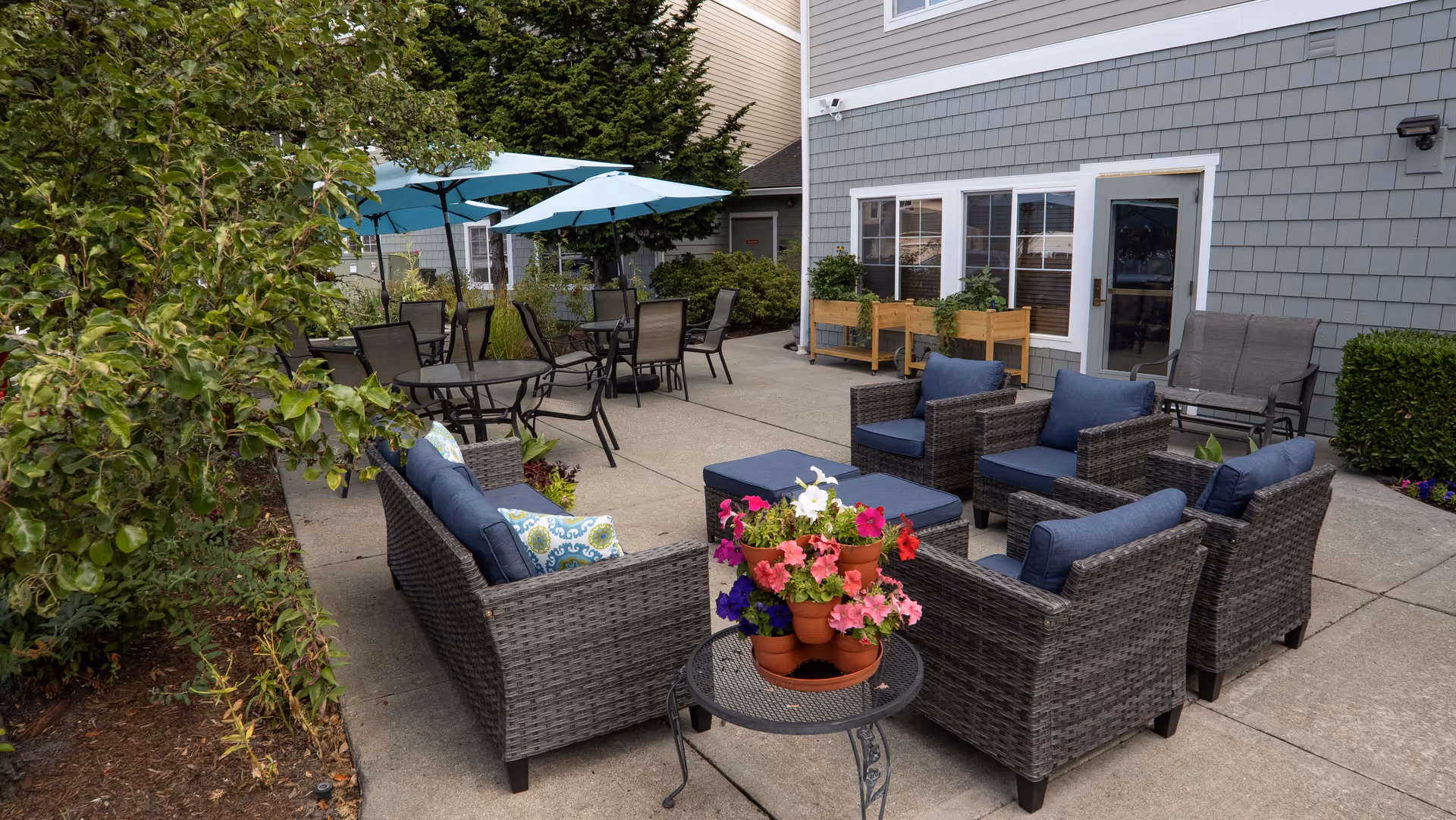 Outdoor patio area at Arbor Village Senior Living featuring wicker seating with blue cushions arranged around a small table with potted flowers, several round tables with chairs and blue umbrellas, and greenery along the side of the building.