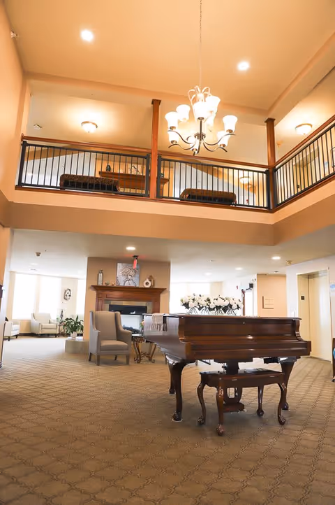 Spacious senior living facility common area with a grand piano in the center, a chandelier hanging from the ceiling, comfortable armchairs, a fireplace, and a second-floor balcony with railings overlooking the area.