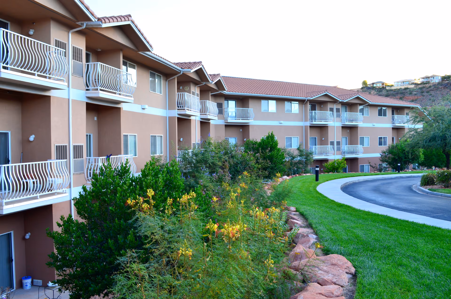 Exterior view of a multi-story assisted living facility with beige walls and red-tiled roofs. The building features multiple balconies with white railings. In front of the building, there is a landscaped area with green grass, bushes, and flowering plants alongside a curved driveway.
