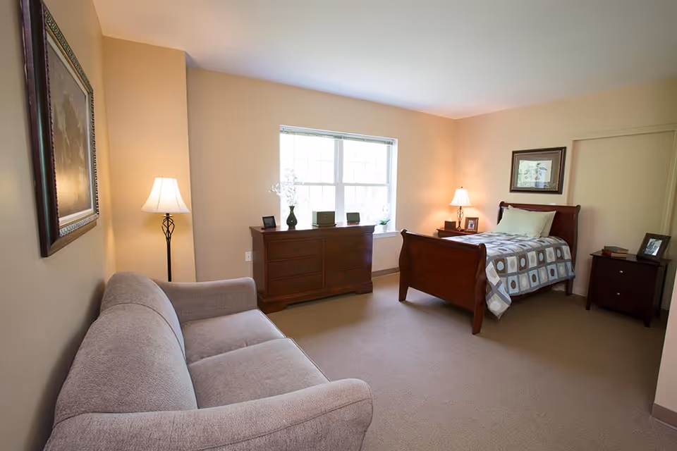 A cozy bedroom in a senior living facility featuring a single wooden bed with a patterned quilt, a wooden dresser under a window, two bedside tables with lamps, a gray upholstered sofa, and framed artwork on the walls. The room is softly lit with natural light coming through the window.