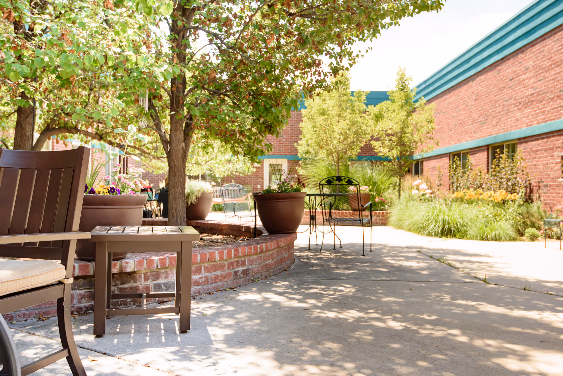 Sunlit outdoor courtyard with patio chairs, potted plants, trees, and a red brick building.