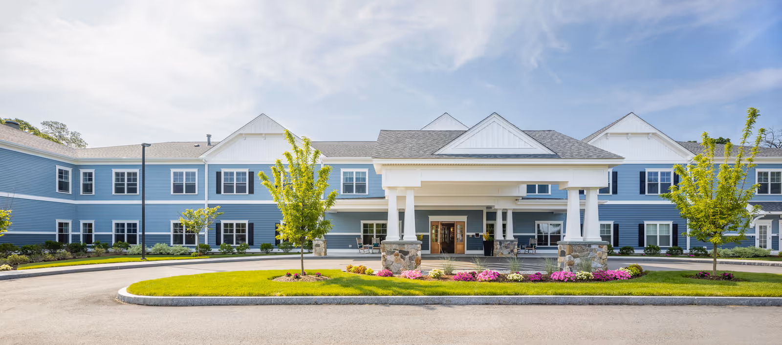 Front exterior view of The Branches of Marlboro facility, a two-story building with blue siding, white trim, and a covered entrance with stone pillars. The driveway curves around a landscaped island with green grass, small trees, and pink flowers under a partly cloudy sky.