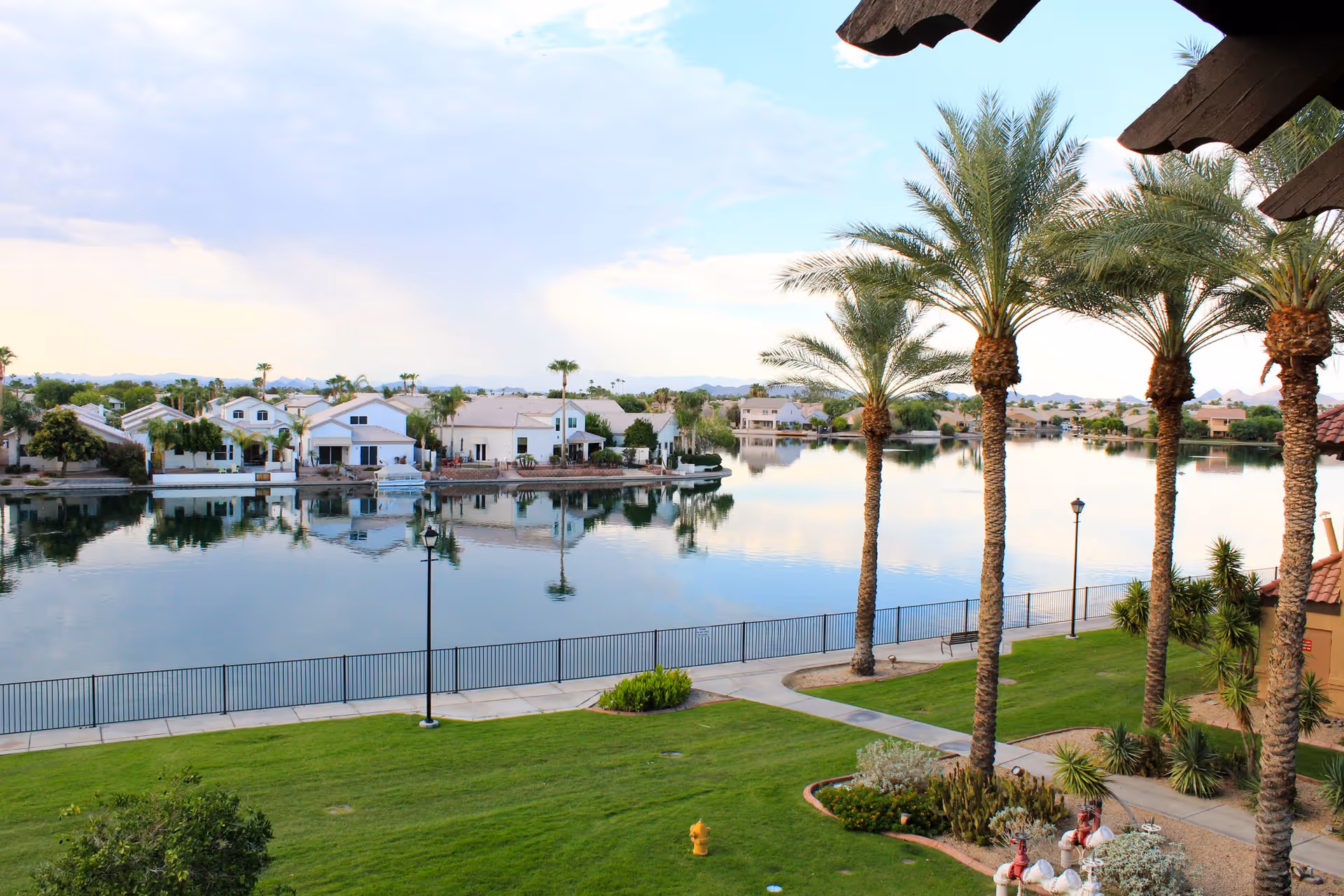 View of a serene lake with houses along the opposite shore, palm trees lining a grassy area with pathways and a black metal fence by the water, under a partly cloudy sky.
