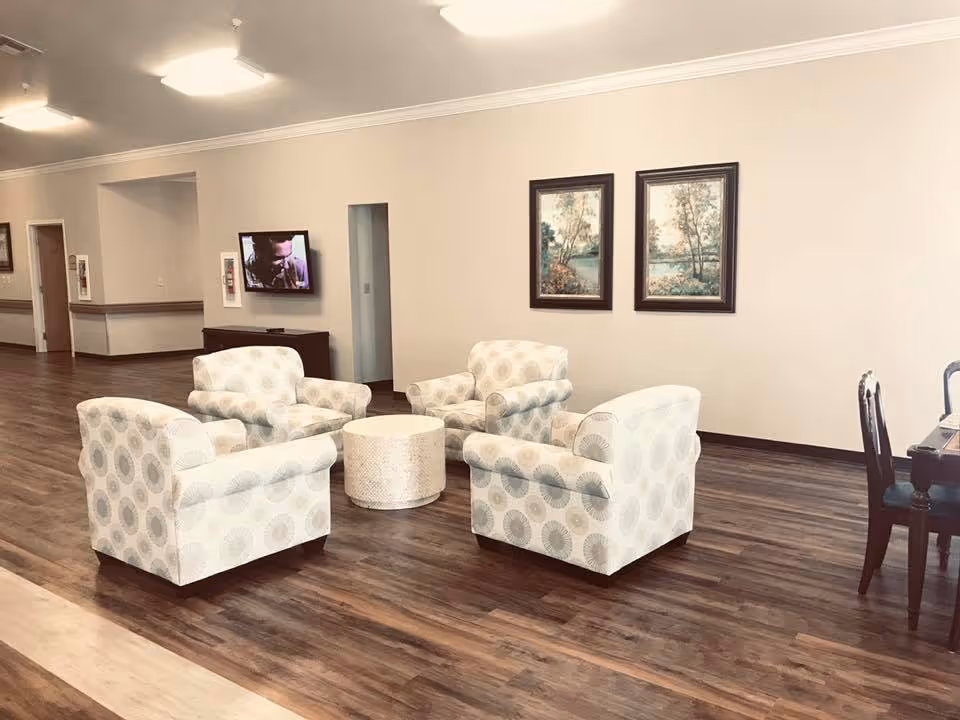A spacious common area with four patterned armchairs arranged around a round white coffee table on a wooden floor. The walls are light-colored with two framed landscape paintings hanging. A flat-screen TV is mounted on the far wall, and there is a wooden cabinet beneath it. To the right, part of a dining table with chairs is visible.