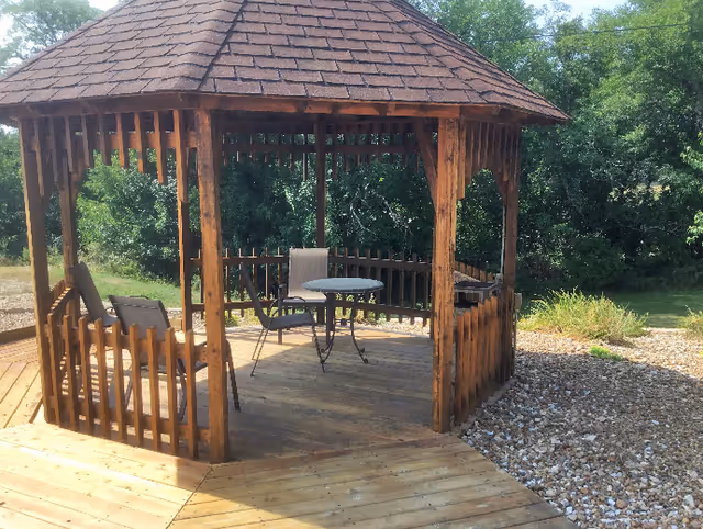 A wooden gazebo with a shingled roof situated on a wooden deck. Inside the gazebo, there is a round metal table and three chairs. Surrounding the gazebo are trees and greenery, with a gravel area adjacent to the deck.
