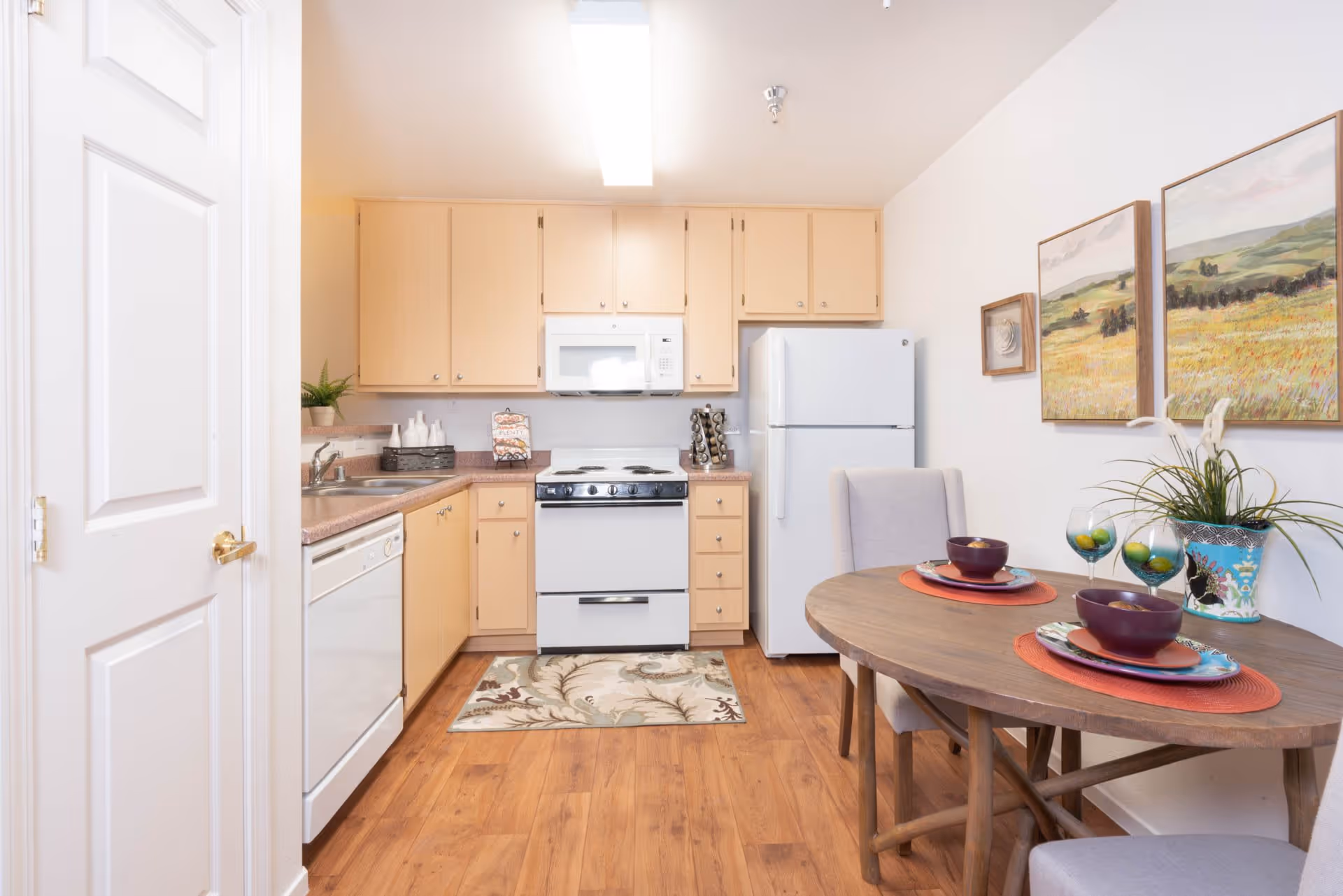 A small kitchen and dining area with light wood cabinets, a white refrigerator, stove, microwave, and dishwasher. The dining table is set with two place settings including bowls, plates, and glasses. There are two chairs around the wooden oval table, and three landscape paintings hang on the wall above the table. The floor is wood, and there is a small rug in front of the stove.