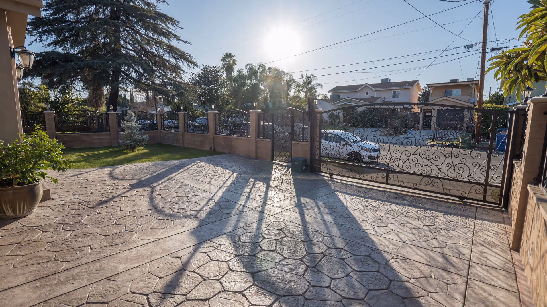 Sunlit gated front patio and driveway with decorative stamped concrete, potted plants, and neighboring houses beyond the gate.