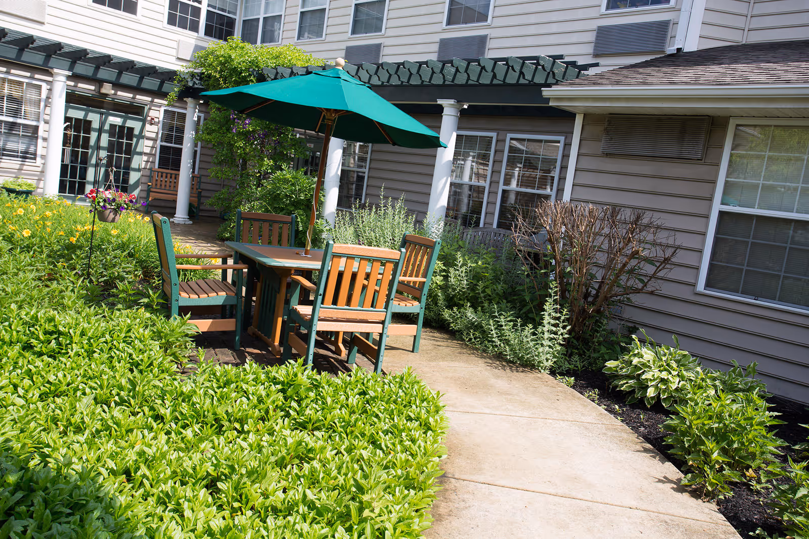Outdoor patio area at Dresher Estates by Priority Life Care featuring a wooden table with four chairs and a green umbrella. The patio is surrounded by lush green plants and flowers, with the building's beige siding and windows in the background.