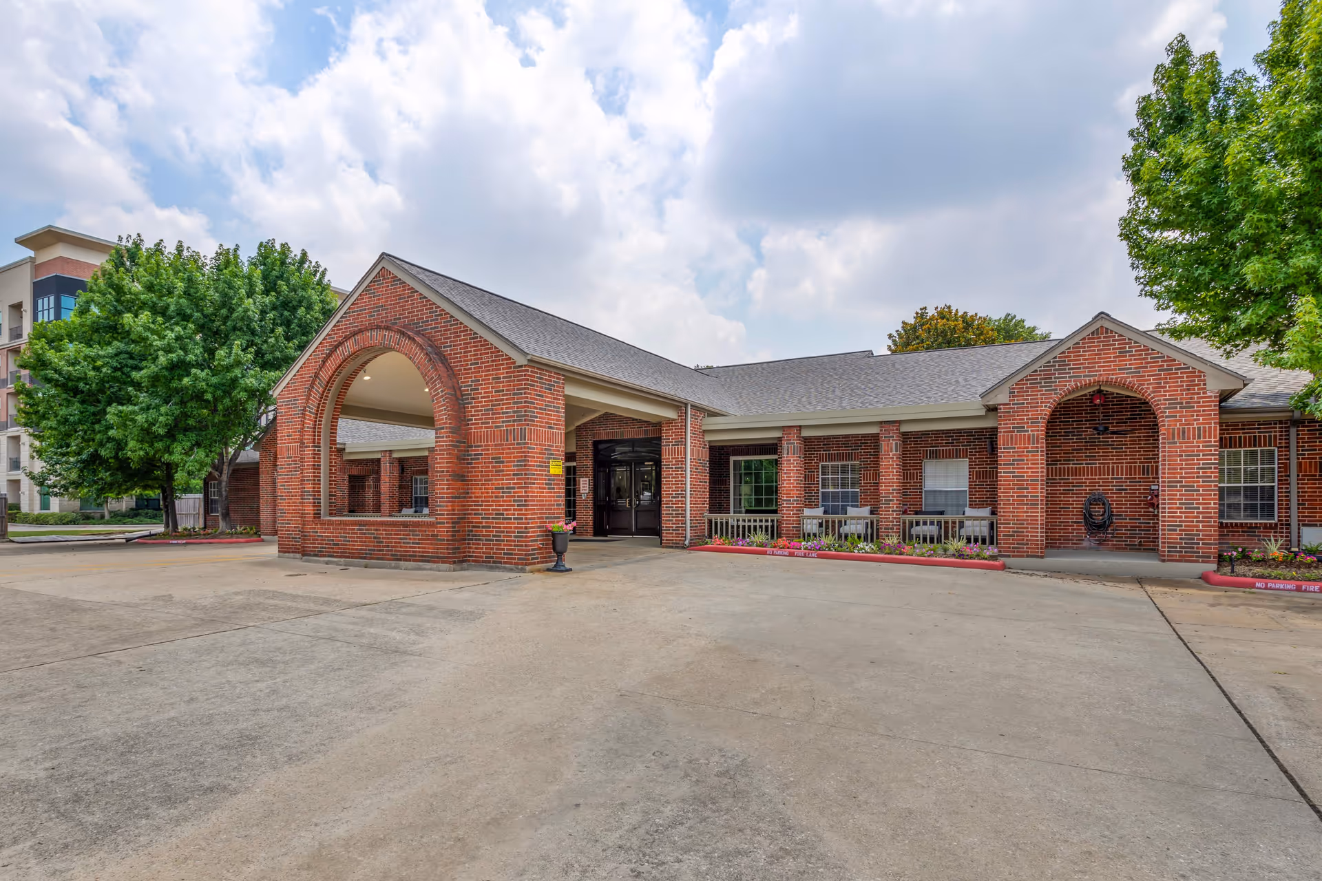 Exterior view of a single-story brick building with arched entryways and a covered driveway. The building is surrounded by trees and has a concrete driveway in front. The sky is partly cloudy.