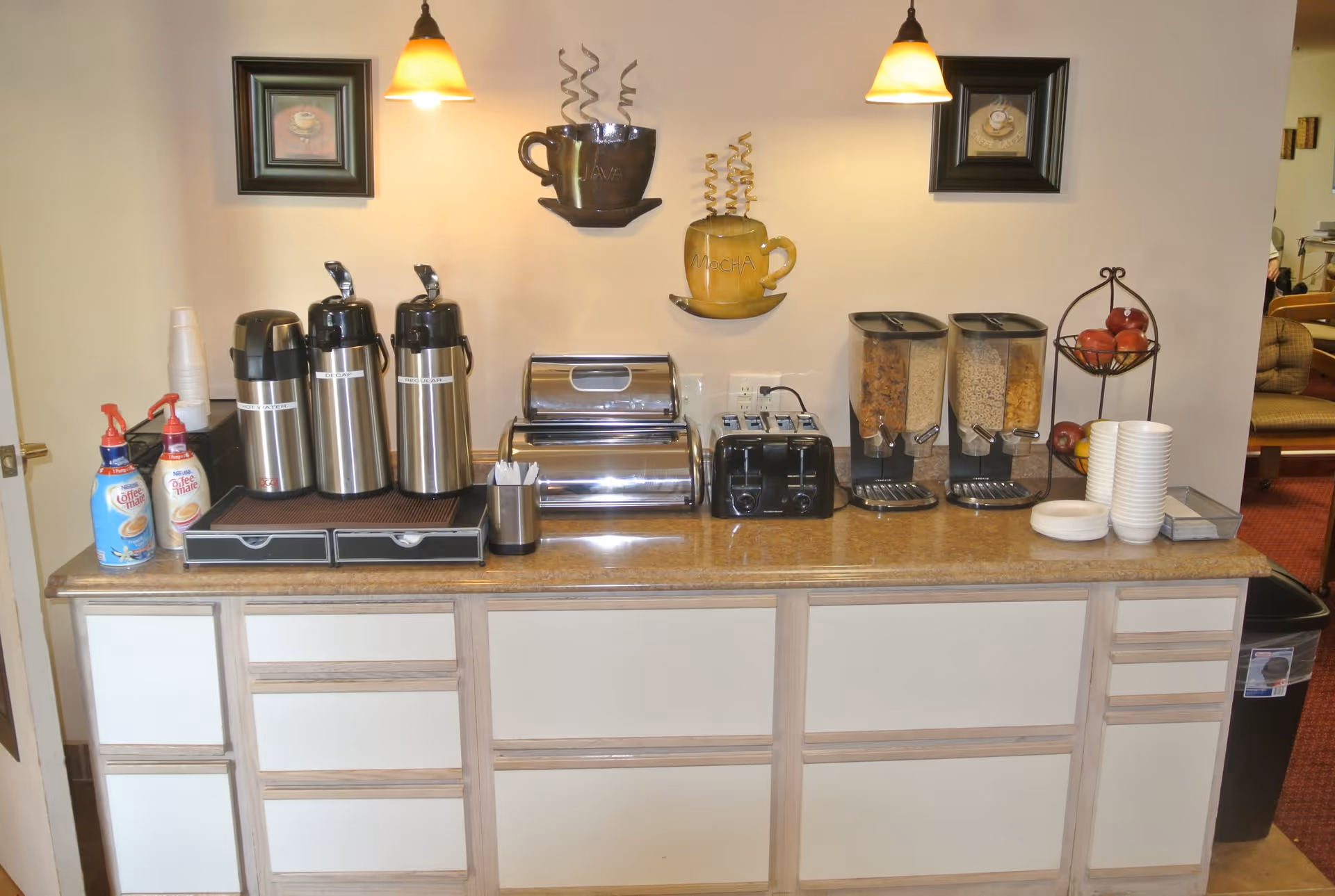 A countertop with coffee dispensers, creamers, a toaster, cereal dispensers, bowls, plates, and apples arranged neatly. Above the counter are two wall-mounted lights and decorative coffee cup wall art labeled 'JAVA' and 'MOCHA'.
