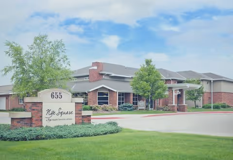 Exterior view of Nye Square, a single-story brick building with a well-maintained lawn and trees under a partly cloudy sky. A sign in front displays the address 655 and the name Nye Square along with 'Life Health Services Campus'.