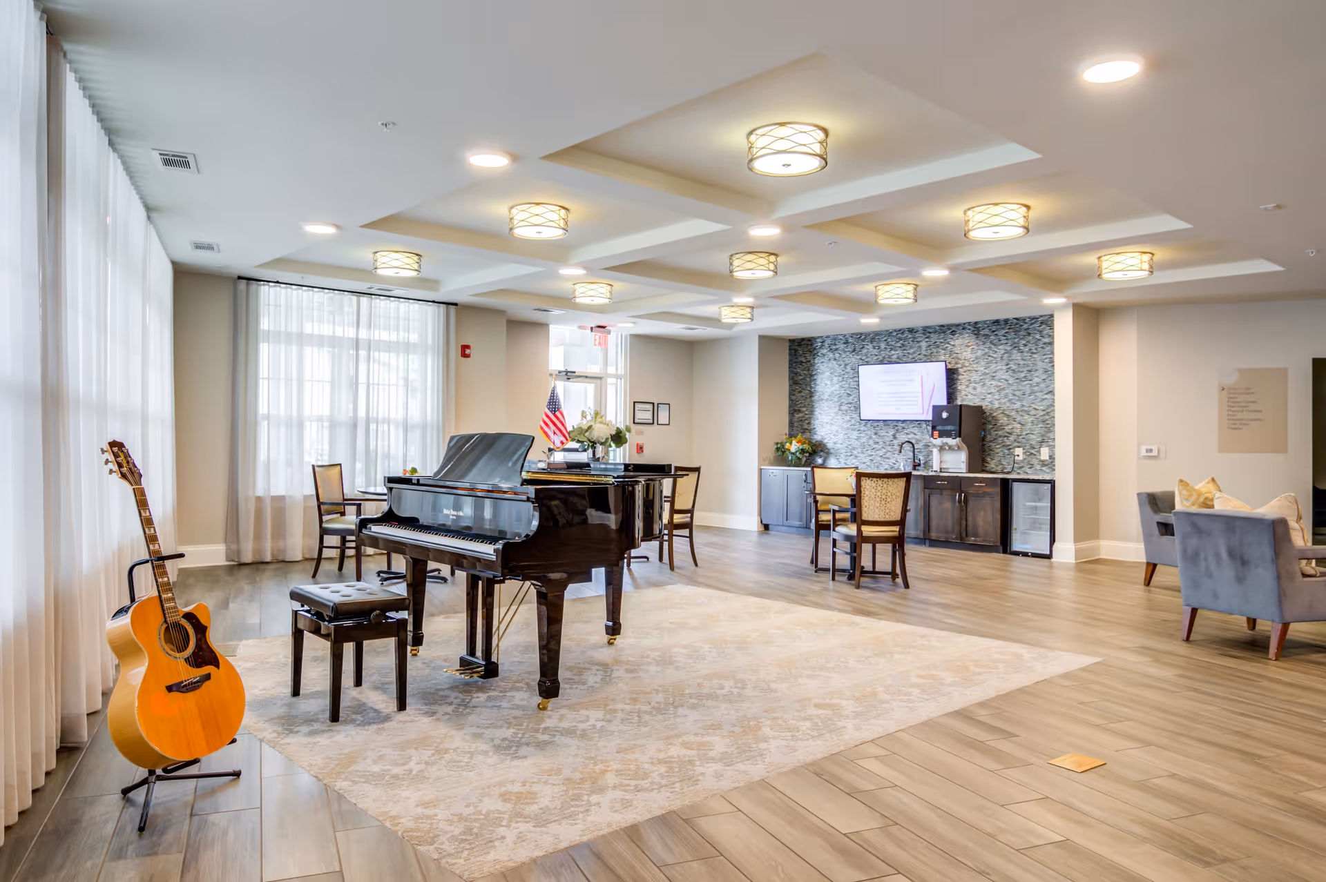 A spacious, well-lit common area with a grand piano and a guitar on a stand near large windows with sheer curtains. The room features a light-colored rug, several chairs and tables, a wall-mounted TV, and a kitchenette area with cabinets and a water dispenser. The ceiling has recessed lighting fixtures.