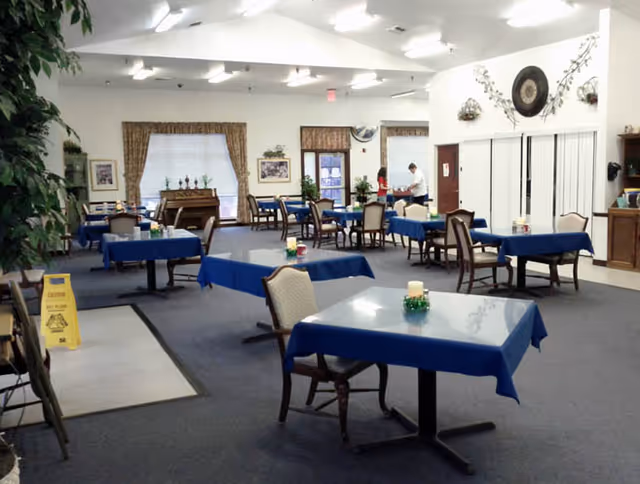 A spacious dining room with multiple tables covered in blue tablecloths, each with a candle centerpiece. Several chairs are arranged around the tables. The room has large windows with curtains, framed pictures on the walls, and a piano in the background. Two people are seen near the back of the room, one standing and one sitting. The floor is carpeted, and there is a caution wet floor sign near the entrance.