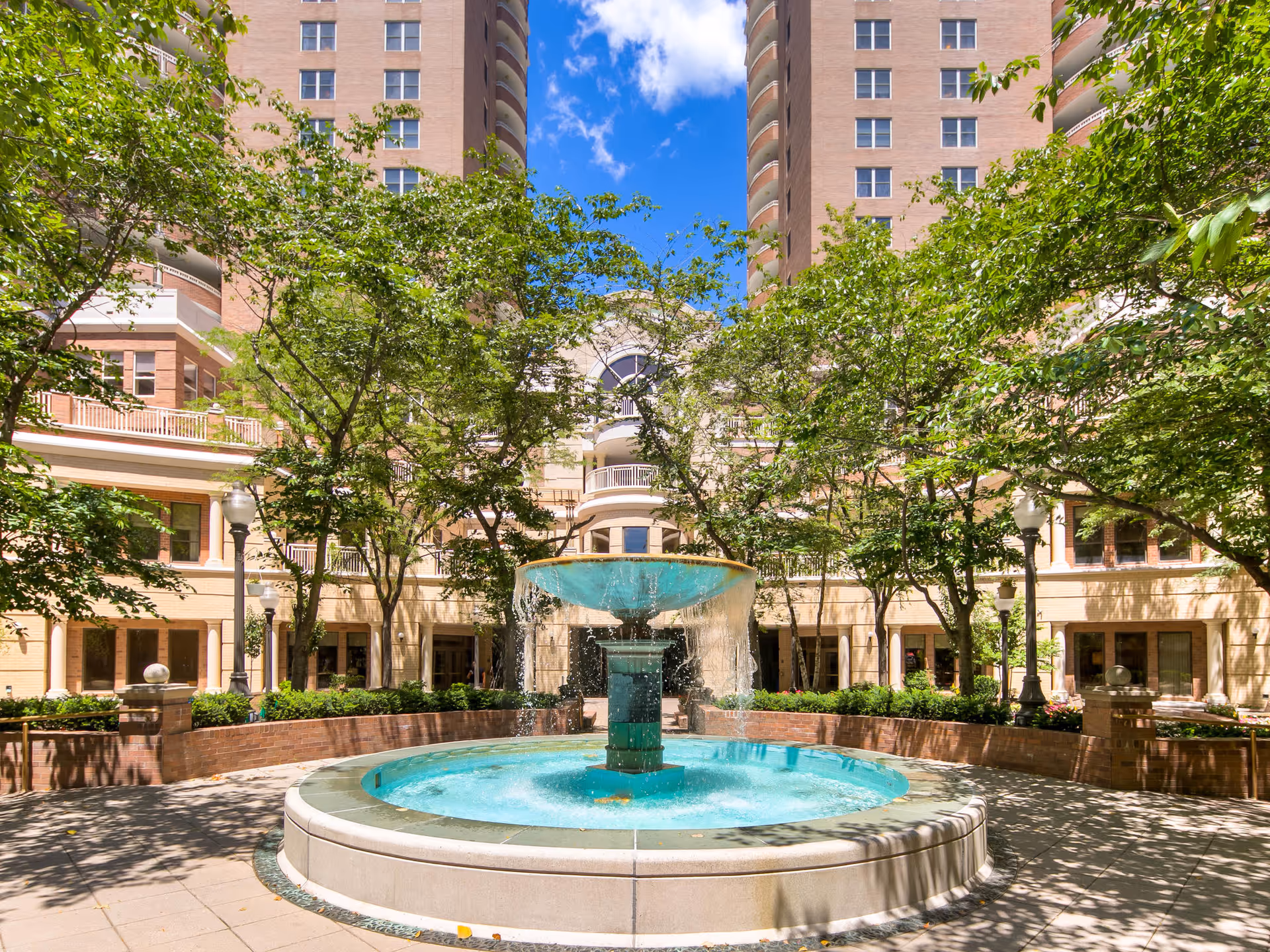Outdoor courtyard area at The Jefferson featuring a large circular water fountain in the center surrounded by trees, brick planters, and tall residential buildings in the background under a clear blue sky.