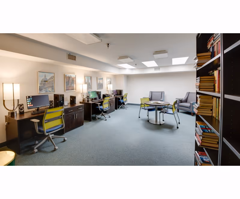 A well-lit room with a row of desks along one wall, each desk equipped with a computer, speakers, and a green office chair. The opposite side of the room has a round table with four green chairs and two gray armchairs. A bookshelf filled with books is visible on the right side. The room has carpeted flooring, framed artwork on the walls, and ceiling lights.