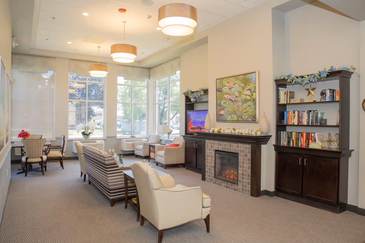 A bright and cozy living room area in an assisted living facility featuring large windows with natural light, a fireplace with decorative items on the mantle, a bookshelf filled with books and decorations, comfortable armchairs and a striped sofa arranged around a coffee table, and a small dining table with chairs near the windows.