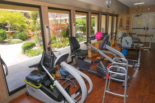 Indoor exercise room with various fitness equipment including recumbent bikes and a walker, large windows showing a garden outside with greenery and a fountain.
