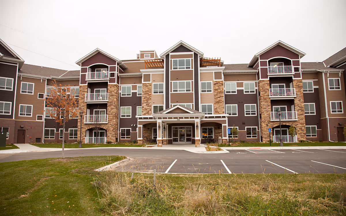 Front exterior view of a multi-story senior living facility building with a covered entrance, balconies, and a parking lot in front. The building features a combination of brown siding and stone accents.