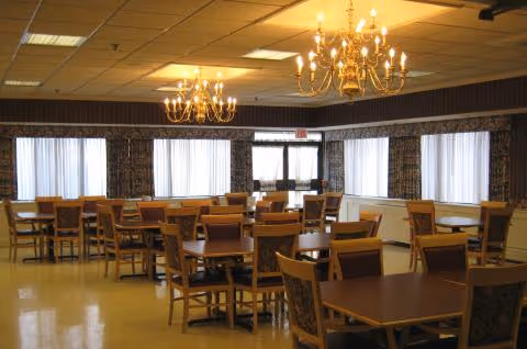 A spacious dining room with multiple wooden tables and chairs arranged neatly. The room features large windows with sheer curtains allowing natural light to enter. Two ornate chandeliers hang from the ceiling, providing warm lighting. The floor is a polished light yellow color.