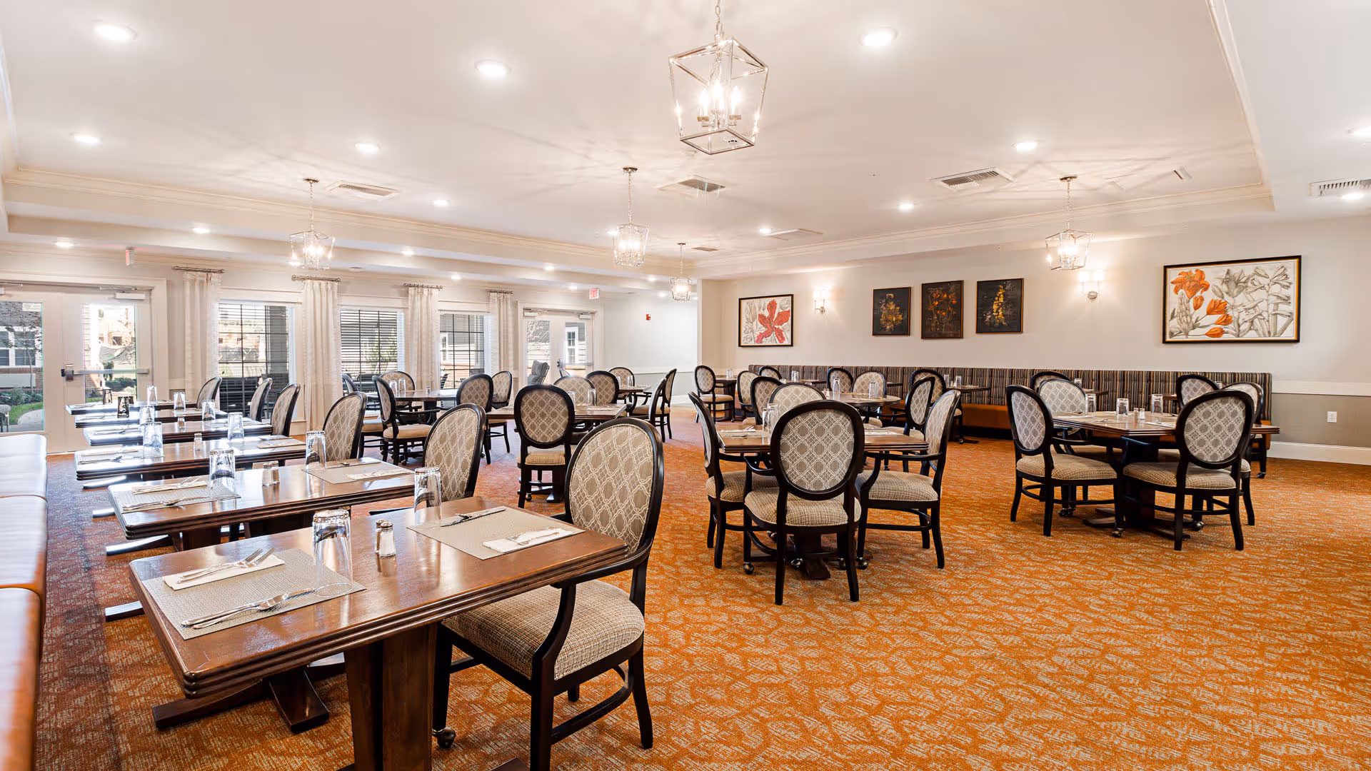 A spacious dining room with multiple wooden tables and cushioned chairs arranged neatly. Each table is set with placemats, silverware, and glasses. The room has large windows with curtains allowing natural light to enter. The walls are decorated with framed floral artwork, and the ceiling features modern light fixtures.