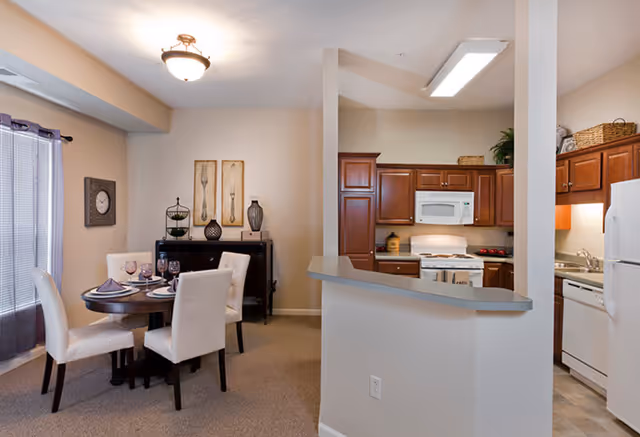 Open-concept dining area with a round table and four upholstered chairs adjacent to a kitchen with wood cabinets and white appliances.