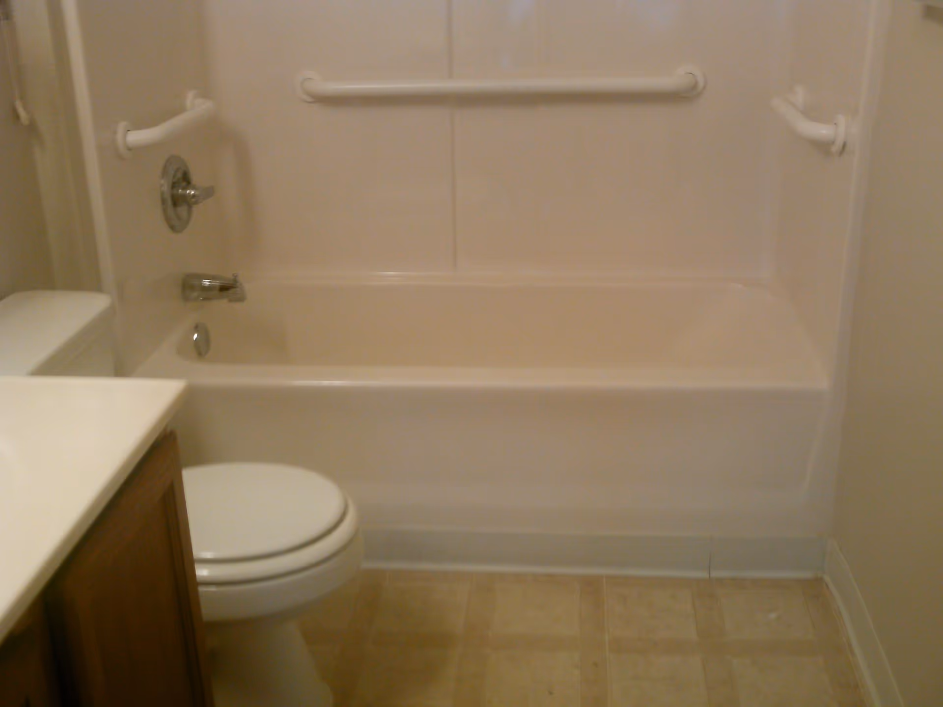 A small bathroom featuring a white bathtub with grab bars on the walls, a toilet, and a vanity with a countertop. The floor has beige tiles.