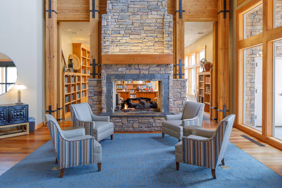 Cozy communal lounge with a central double-sided stone fireplace surrounded by four armchairs on a blue rug, with bookshelves and large windows in the background.